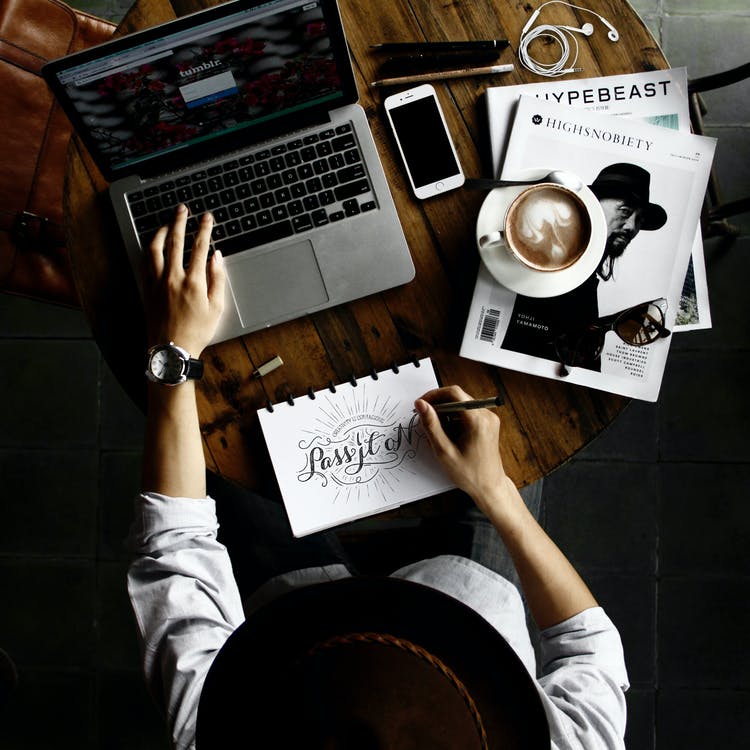 La imagen muestra una persona sentada haciendo varias cosas a la vez. La persona está escribiendo en su cuaderno, buscando información en el ordenador. En la mesa está el móvil y el café a la derecha. 