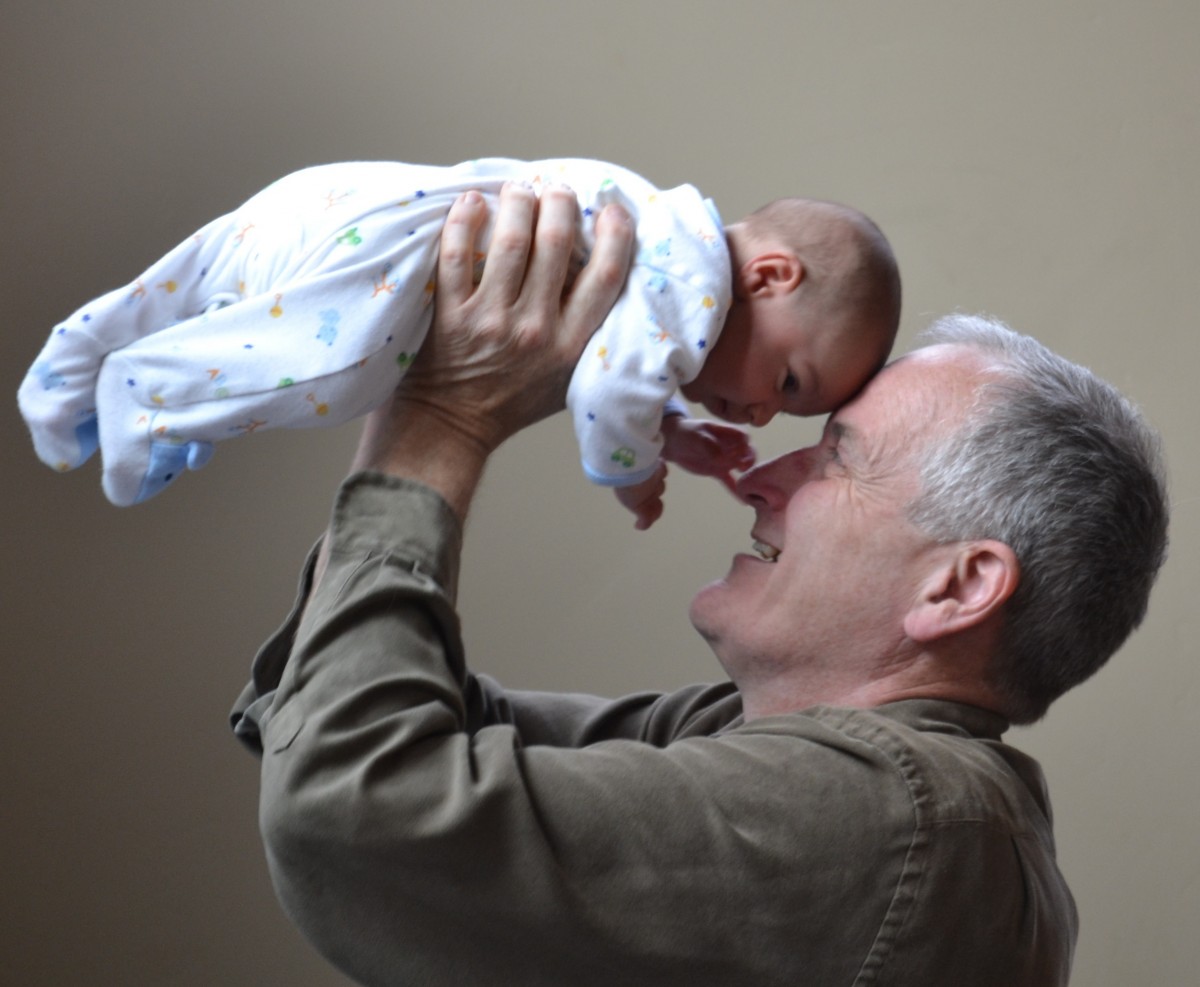 La imagen muestra a un abuelo levantando a su nieto en brazos y juntando su frente con la del bebé.