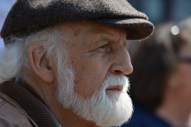 La imagen muestra el perfil de un hombre mayor con barba blanca con una gorra plana en la cabeza.