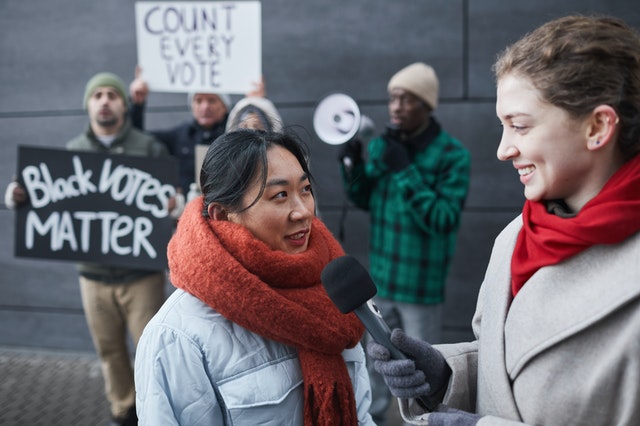 La imagen muestra a una reportera entrevistando a una mujer de origen asiático y un grupo de jóvenes detrás de ellas portando carteles a modo de manifestación.