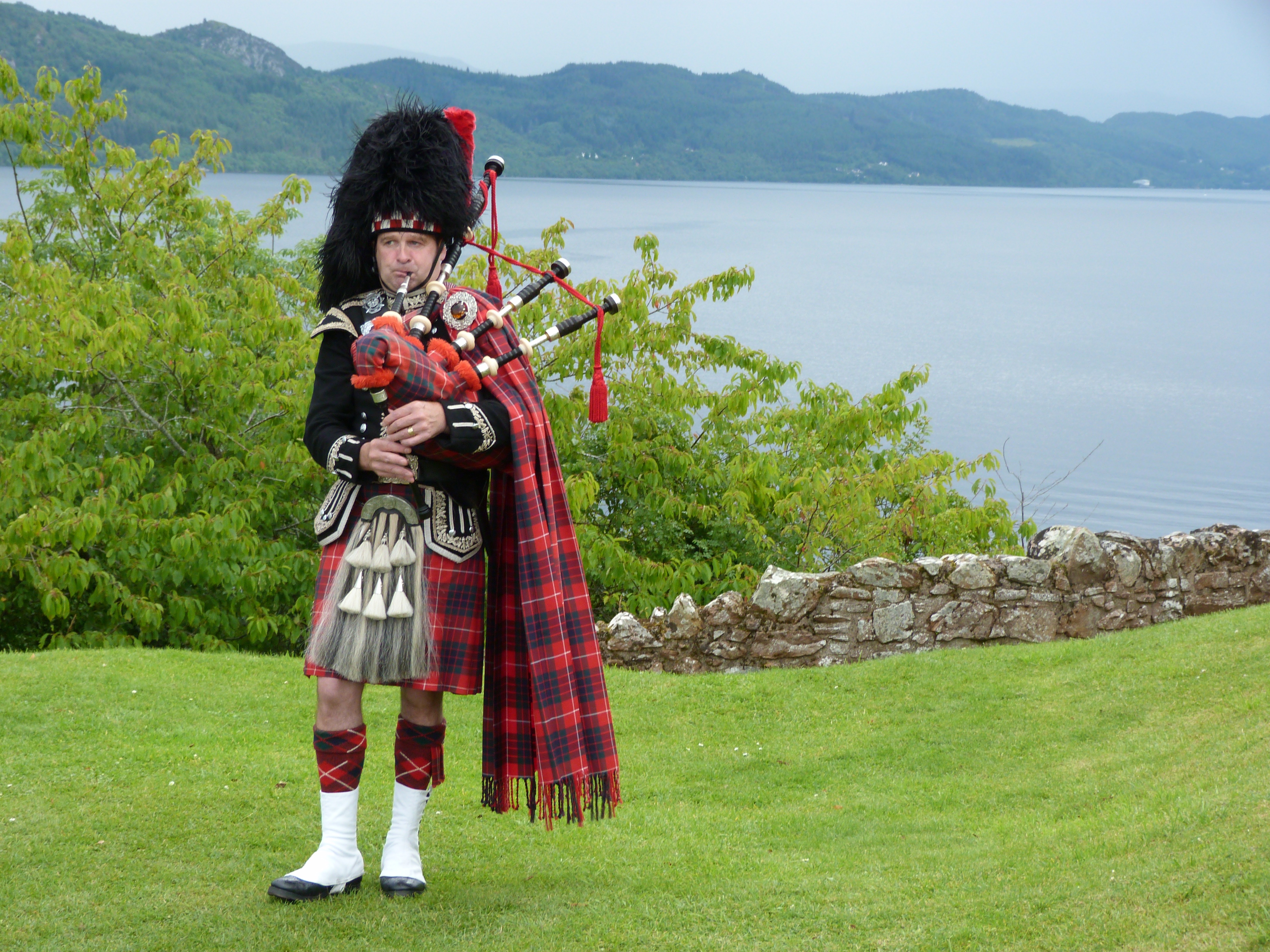 Imagen de un escocés, vestido con la falda típica escocesa tocando una gaita. Se ve el cuerpo entero de la cabeza a los pies.