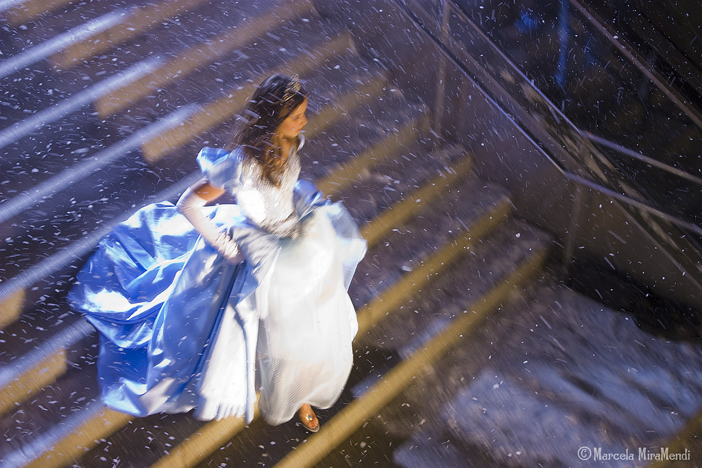 Imagen tomada desde arriba, se ve a una chica vestida con un vestido de gala azul y blanco, bajando rápidamente una escaleras. Lleva puestos unos zapatos de cristal