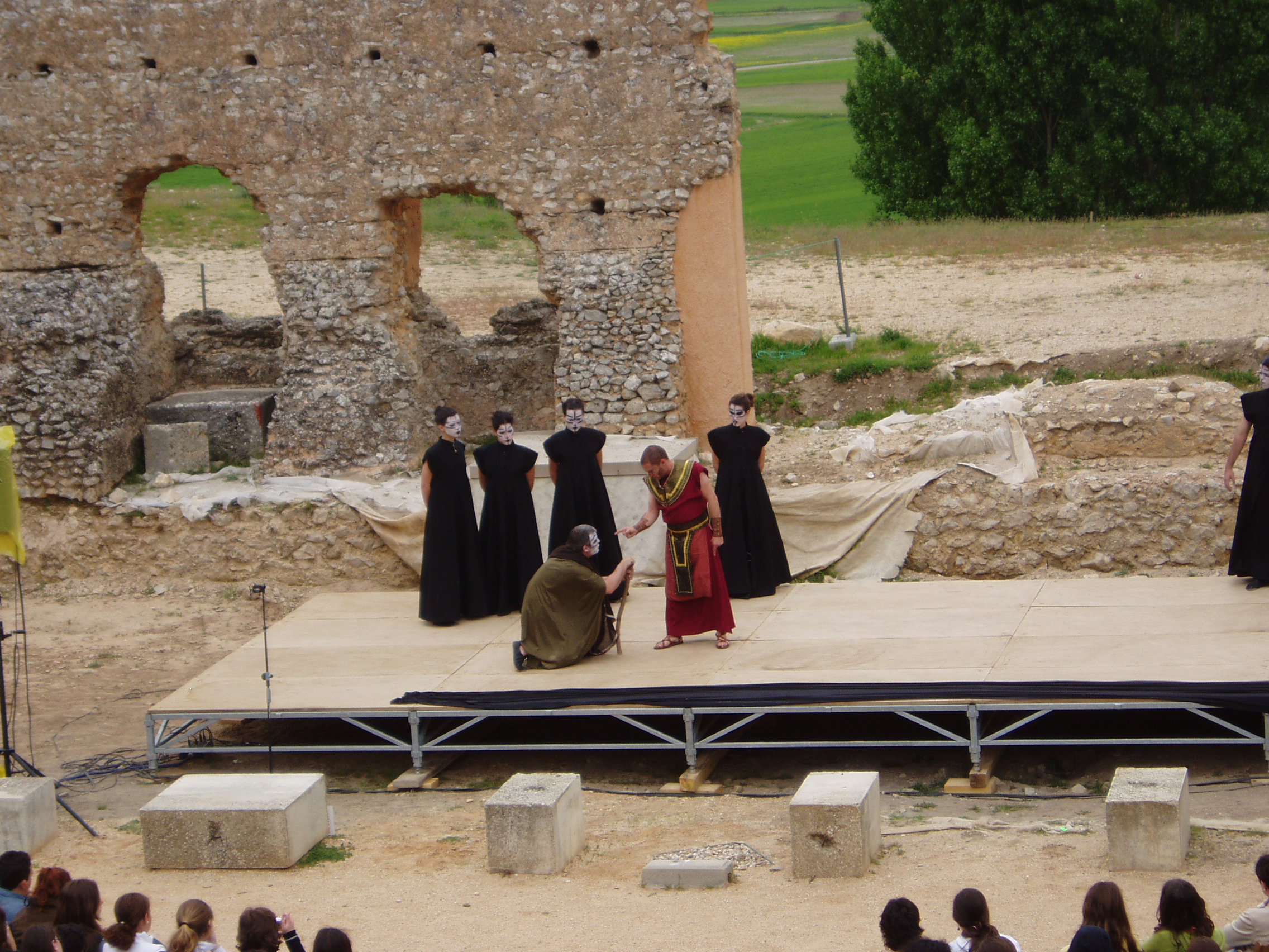 Escena teatral donde aparece un escenario exterior con poca altura. En él aparecen 6 personas, cuatro de ellas vestidas con traje negro y la cara pintada de negro. Se puede apreciar a los espectadores.