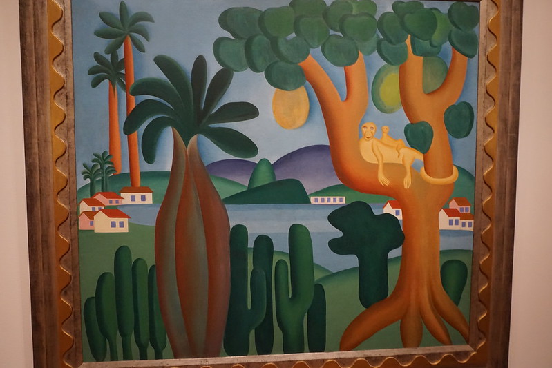 Cuadro colorido que muestra un paisaje tropical con colores verdes, palmeras, casas blancas y una pequeña laguna