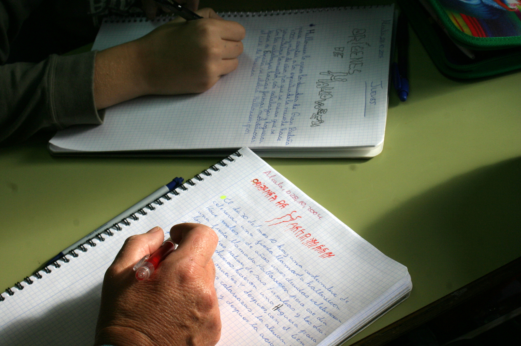 La imagen muestra las manos de dos personas sobre una mesa escribiendo sobre sendas libretas