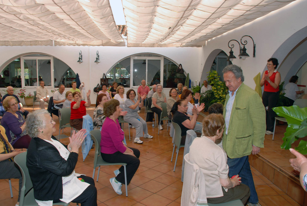 Esta imagen muestra a un grupo de persona s mayores en un salón sentadas y aplaudiendo a un hombre