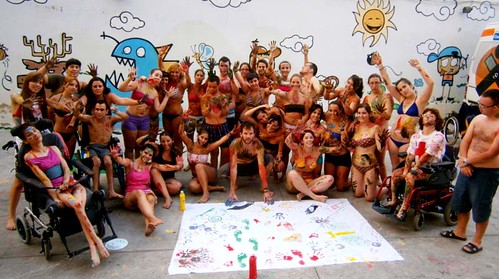 Esta imagen muestra un grupo de personas manchadas de pintura con un mural con sus manos estampadas en el suelo
