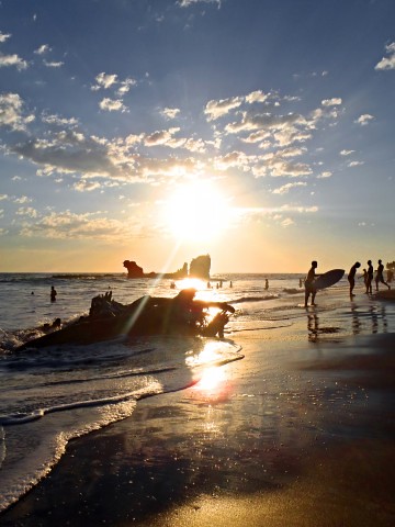Esta imagen muestra el atardecer en una playa donde hay gente con tablas de surf, gente dentro del mar y otras personas en la orilla.