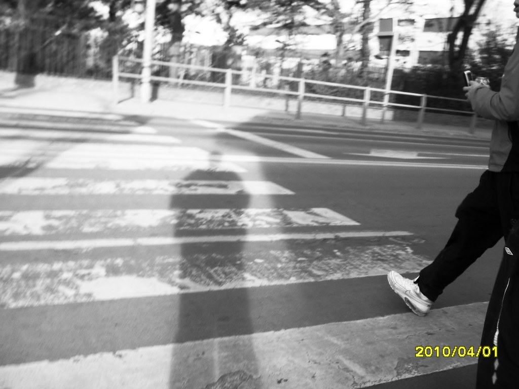 Esta imagen muestra un paso de peatones con una sombra de una persona y unos pies comenzado a caminar 