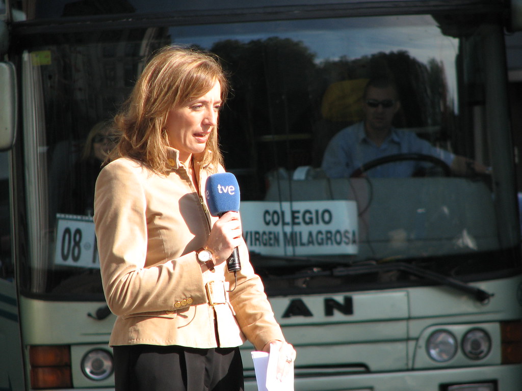 Esta imagen muestra una mujer con un micro en la mano hablando y detrás de ella un autobús