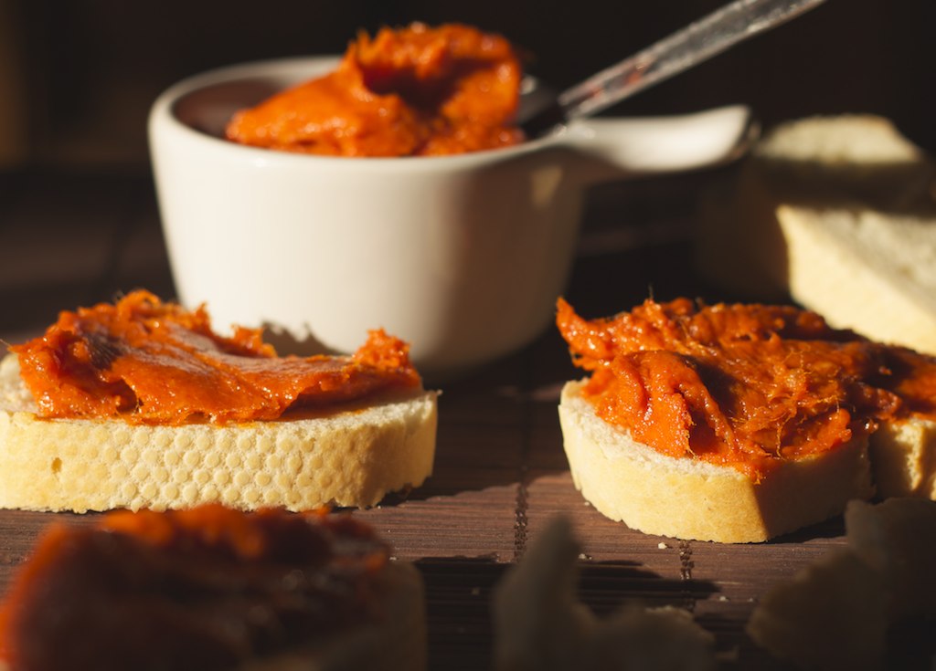 Esta imagen muestra dos rebanadas de pan untadas en una crema naranja hecha con manteca y detrás de ellas un tarro con más crema naranja